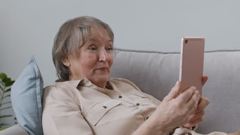 Glückliche-Seniorin,-Die-Einen-Videoanruf-Per-Tablet-Hat,-Während-Sie-Zu-Hause-Auf-Dem-Sofa-Liegt-2