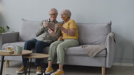 Arabische-Frau-Lehrt-Einen-älteren-Mann,-Tablet-Zu-Benutzen,-Während-Sie-Zu-Hause-Auf-Dem-Sofa-Sitzen