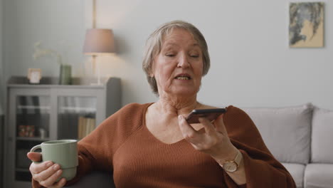 Senior-Woman-Making-Hands-Free-Phone-Call-At-Home-While-Holding-A-Coffee-Cup-Sitting-On-Sofa-At-Home