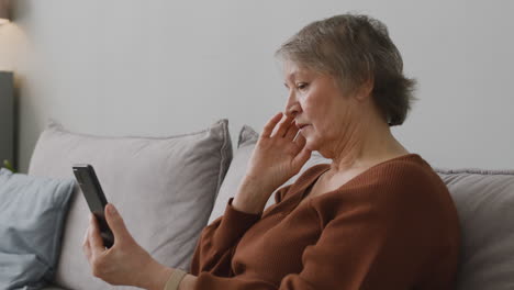 Senior-Woman-Using-Smartphone-While-She-Is-Sitting-On-Sofa-In-Living-Room-At-Home