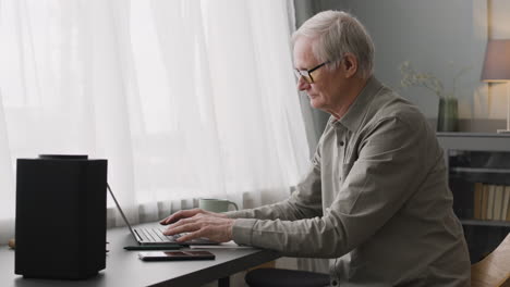 Glücklicher-älterer-Mann,-Der-Am-Laptop-Schreibt,-Während-Er-Am-Schreibtisch-Sitzt