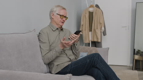 älterer-Mann,-Der-Einen-Videoanruf-Mit-Dem-Smartphone-Tätigt,-Während-Er-Zu-Hause-Auf-Dem-Sofa-Im-Wohnzimmer-Sitzt