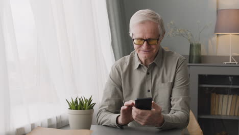 Glücklicher-älterer-Mann-Mit-Smartphone-Am-Schreibtisch-Im-Wohnzimmer-Zu-Hause-Sitzen