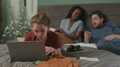 Mädchen-Mit-Laptop-Auf-Dem-Bett-Liegend,-Während-Ihre-Mitbewohner-Im-Hintergrund-Sprechen