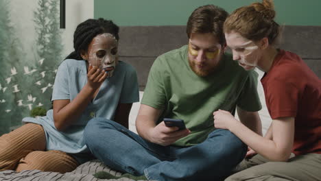 Three-Rommates-With-Facial-Mask-Watching-Smartphone-Sitting-On-The-Bed-In-Bedroom