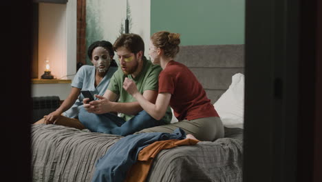 Three-Rommates-With-Facial-Mask-Watching-Smartphone-Sitting-On-The-Bed-In-Bedroom-1