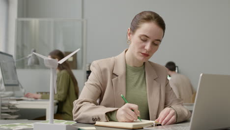 Mujer-Usando-Laptop-Y-Escribiendo-Notas-Sentada-En-La-Mesa-Con-Modelo-De-Molino-De-Viento-En-La-Oficina