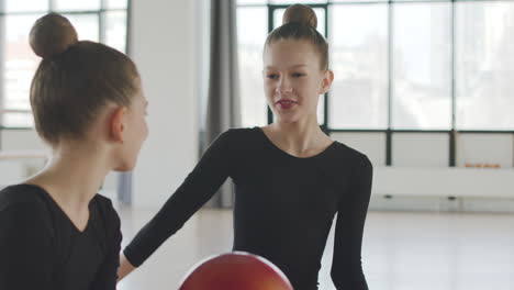 Gymnastic-Blonde-Girl-Playing-With-A-Band-While-Talking-With-Her-Friend-Before-Starting-Ballet-Class