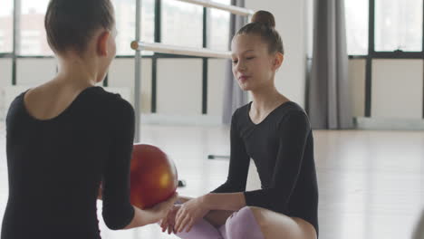 Two-Gymnastic-Blonde-Girls-Talking-While-Playing-With-A-Ball-Sitting-On-The-Floor-Before-Starting-Ballet-Class