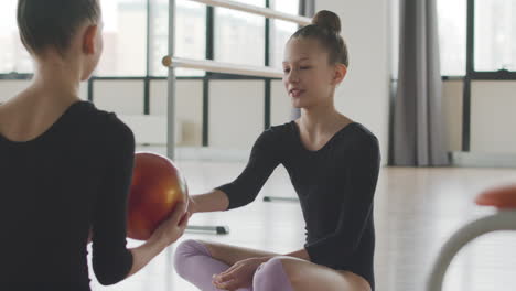 Two-Gymnastic-Blonde-Girls-Talking-While-Playing-With-A-Ball-Sitting-On-The-Floor-Before-Starting-Ballet-Class-1