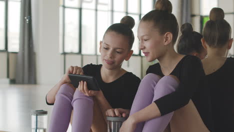 Two-Gymnastic-Blonde-Girls-Using-Smartphone-Sitting-On-The-Floor-Before-Starting-Ballet-Class