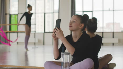 Gymnastic-Blonde-Girl-Sitting-On-The-Floor-And-Taking-Photo-Of-Her-Friend-Who-Is-Dancing-Holding-A-Band-1