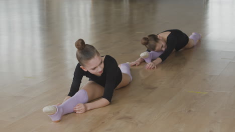 Two-Gymnastic-Blonde-Girls-With-Legs-Outstretched-Rehearsing-A-Ballet-Move-On-The-Floor