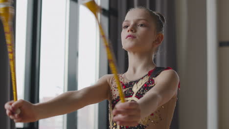 Focused-Young-Girl-In-Leotard-Practising-Rhythmic-Gymnastics-With-Clubs-In-A-Studio