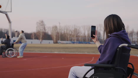 Behinderte-Frau-Im-Rollstuhl-Nimmt-Mit-Smartphone-Zu-Ihren-Freunden-Auf,-Die-Basketball-Spielen