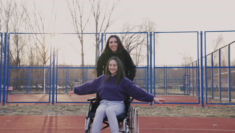 Junge-Frau-Läuft,-Während-Sie-Ihren-Behinderten-Freund-Im-Rollstuhl-Schiebt-Und-Gemeinsam-Spaß-Auf-Dem-Basketballplatz-Hat