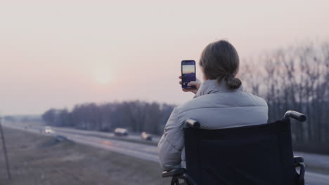 Rear-View-Of-Disabled-Woman-In-Wheelchair-Recording-The-Landscape-At-Sunset
