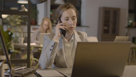 Businesswoman-Talking-On-Mobile-Phone-While-Working-On-Laptop-Computer-In-The-Office