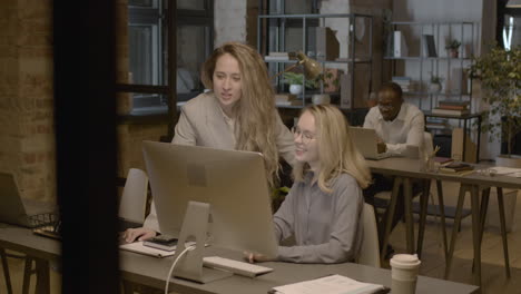 Two-Female-Employees-Looking-Something-At-Computer-And-Talking-Together-In-The-Office