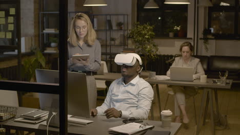 Amerikanischer-Mann-Sitzt-Am-Schreibtisch-Im-Büro-Und-Trägt-Eine-Virtual-reality-brille,-Während-Eine-Angestellte-Ihn-Mit-Einem-Tablet-Steuert