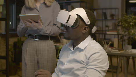 Nahaufnahme-Eines-Amerikanischen-Mannes,-Der-Am-Schreibtisch-Im-Büro-Sitzt-Und-Eine-Virtual-Reality-Brille-Trägt,-Während-Eine-Mitarbeiterin-Ihn-Mit-Einem-Tablet-Steuert