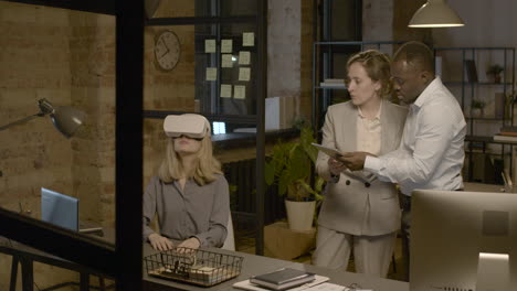 Frau-Am-Schreibtisch-Im-Büro-Mit-Virtual-Reality-Brille,-Während-Eine-Frau-Und-Ein-Mann-Sie-Mit-Einem-Tablet-Steuern