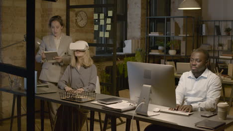 Mitarbeiterin-Am-Schreibtisch-Im-Büro-Mit-Virtual-Reality-Brille,-Während-Ihr-Kollege-Sie-Mit-Tablet-Steuert