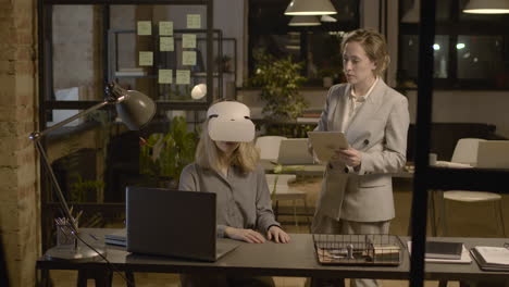 Woman-Emplooyee-At-Desk-In-The-Office-Wearing-Virtual-Reality-Glasses-While-Her-Coworker-Is-Controlling-Her-With-Tablet-2