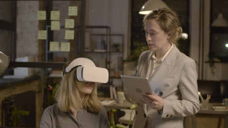 Woman-Emplooyee-At-Desk-In-The-Office-Wearing-Virtual-Reality-Glasses-While-Her-Coworker-Is-Controlling-Her-With-Tablet-3