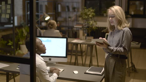 Rear-View-Of-American-Man-Sitting-At-Desk-While-Talking-With-Female-Coworker-Who-Is-Holding-A-Tablet-In-The-Office