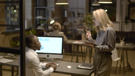 Rear-View-Of-American-Man-Sitting-At-Desk-While-Talking-With-Female-Coworker-Who-Is-Holding-A-Tablet-In-The-Office-1