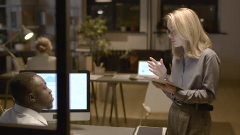 Rear-View-Of-American-Man-Sitting-At-Desk-While-Talking-With-Female-Coworker-Who-Is-Holding-And-Watching-A-Tablet-In-The-Office