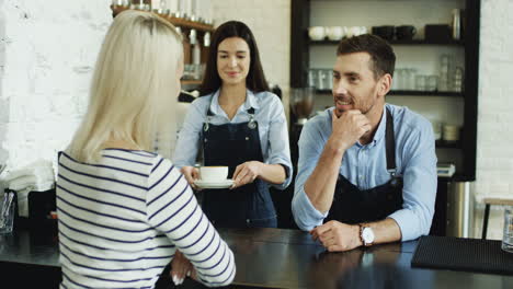 Camarero-Hablando-Con-Una-Mujer-Rubia-En-El-Bar,-Mientras-Una-Camarera-Le-Sirve-Café