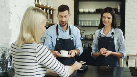Rear-View-Of-Blonde-Woman-Using-Her-Smartphone-To-Pay-A-Coffee-While-The-Waiter-Serving-It-And-The-Waitress-Use-The-Card-Reader