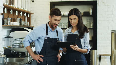 Young-Waitress-Scrolling-And-Tapping-On-The-Device-And-Her-Male-Coworker-Standing-Close-And-Waching-The-Screen-At-The-Cafe