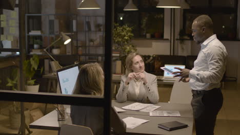 American-Man-Employee-Holding-A-Tablet-And-Explaining-A-Project-To-Female-Coworkers-Who-Are-Sitting-At-Desk-In-The-Office