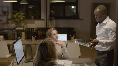 American-Man-Employee-Holding-A-Tablet-And-Explaining-A-Project-To-Female-Coworkers-Who-Are-Sitting-At-Desk-In-The-Office-1