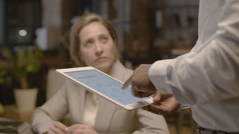Close-Up-View-Of-Unrecognizable-American-Man-Employee-Holding-A-Tablet-And-Explaining-Graphics-To-Female-Coworkers-Who-Are-Sitting-At-Desk-In-The-Office