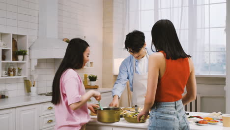 Drei-Japanische-Freunde-Gießen-Beim-Kochen,-Reden-Und-Lachen-In-Der-Küche-Zutaten-In-Die-Pfanne