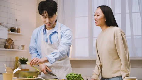 Japanese-Man-Cutting-Ingredients-While-Talking-With-Her-Friend-In-The-Kitchen,-Then-He-Gives-Her-Some-Food-To-Taste