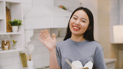 Fröhliches-Japanisches-Mädchen,-Das-Ramen-Zum-Mitnehmen-Isst,-Während-Es-In-Die-Kamera-Schaut-Und-Lächelnd-Winkt