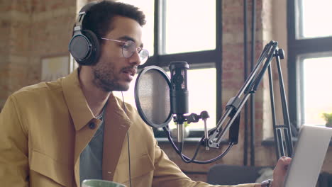 Man-Recording-A-Podcast-Wearing-Eyeglasses-And-Headphones-Talking-Into-A-Microphone-While-Sitting-On-A-Table-With-Laptop