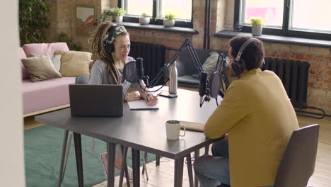 Young-Man-And-Woman-Sitting-At-A-Table-With-Microphones-And-Computer-While-Recording-A-Podcast