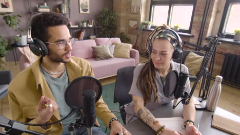 High-Angle-View-Of-Young-Man-And-Woman-Wearing-Headphones-Sitting-At-A-Table-With-Microphones-While-They-Recording-A-Podcast