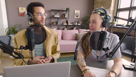 Front-View-Of-Young-Man-And-Woman-Wearing-Headphones-Sitting-At-A-Table-With-Microphones-While-They-Recording-A-Podcast