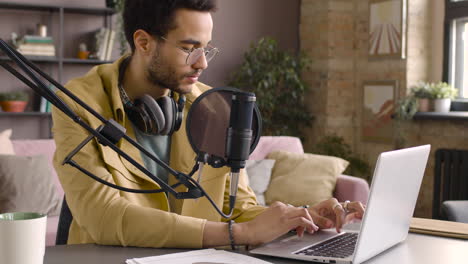 Man-Wearing-Glasses-And-Sitting-At-A-Table-With-Microphone-While-Typewriting-On-Laptop-1