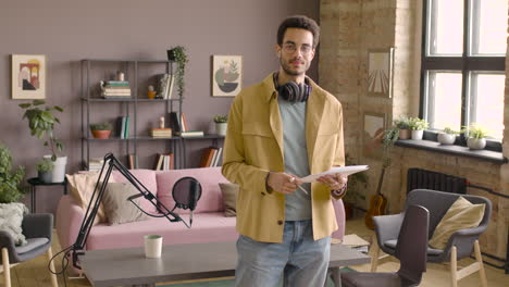 Man-With-Headphones-Standing-Holding-Papers-In-A-Room-With-Microphone-And-Looking-At-Camera