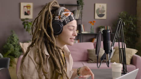 Side-View-Of-Woman-With-Dreadlocks-Recording-A-Podcast-Wearing-Headphones-And-Talking-Into-A-Microphone-Sitting-At-A-Desk