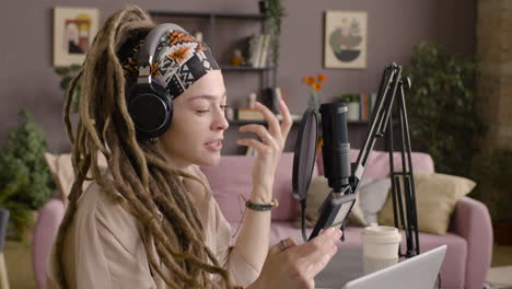 Side-View-Of-Woman-With-Dreadlocks-Recording-A-Podcast-Wearing-Headphones,-Talking-Into-A-Microphone-And-Reading-On-Smartphone-Sitting-At-A-Desk