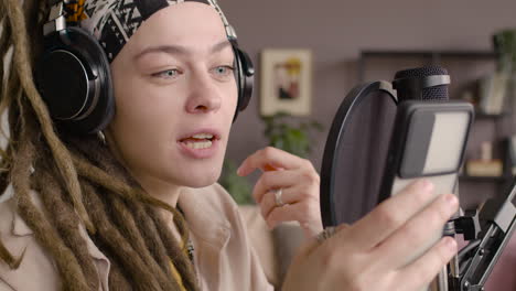 Close-Up-View-Of-Woman-With-Dreadlocks-Recording-A-Podcast-Wearing-Headphones,-Talking-Into-A-Microphone-And-Reading-On-Smartphone-Sitting-At-A-Desk